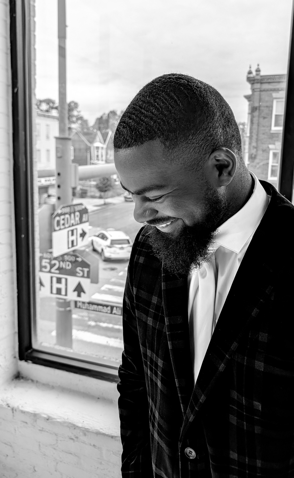 Nasir Leach in a plaid blazer and tie, smiling by a window in a black-and-white portrait